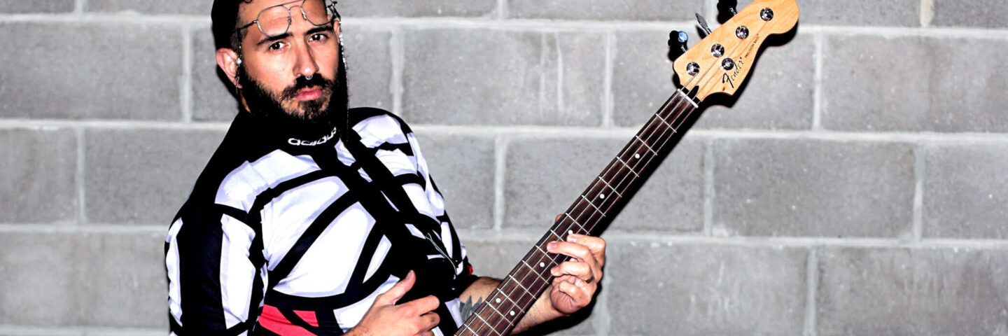 A white person with a dark beard wears glasses, a black baseball cap, a patterned skirt and a black and white sweater. They hold an electric guitar. 