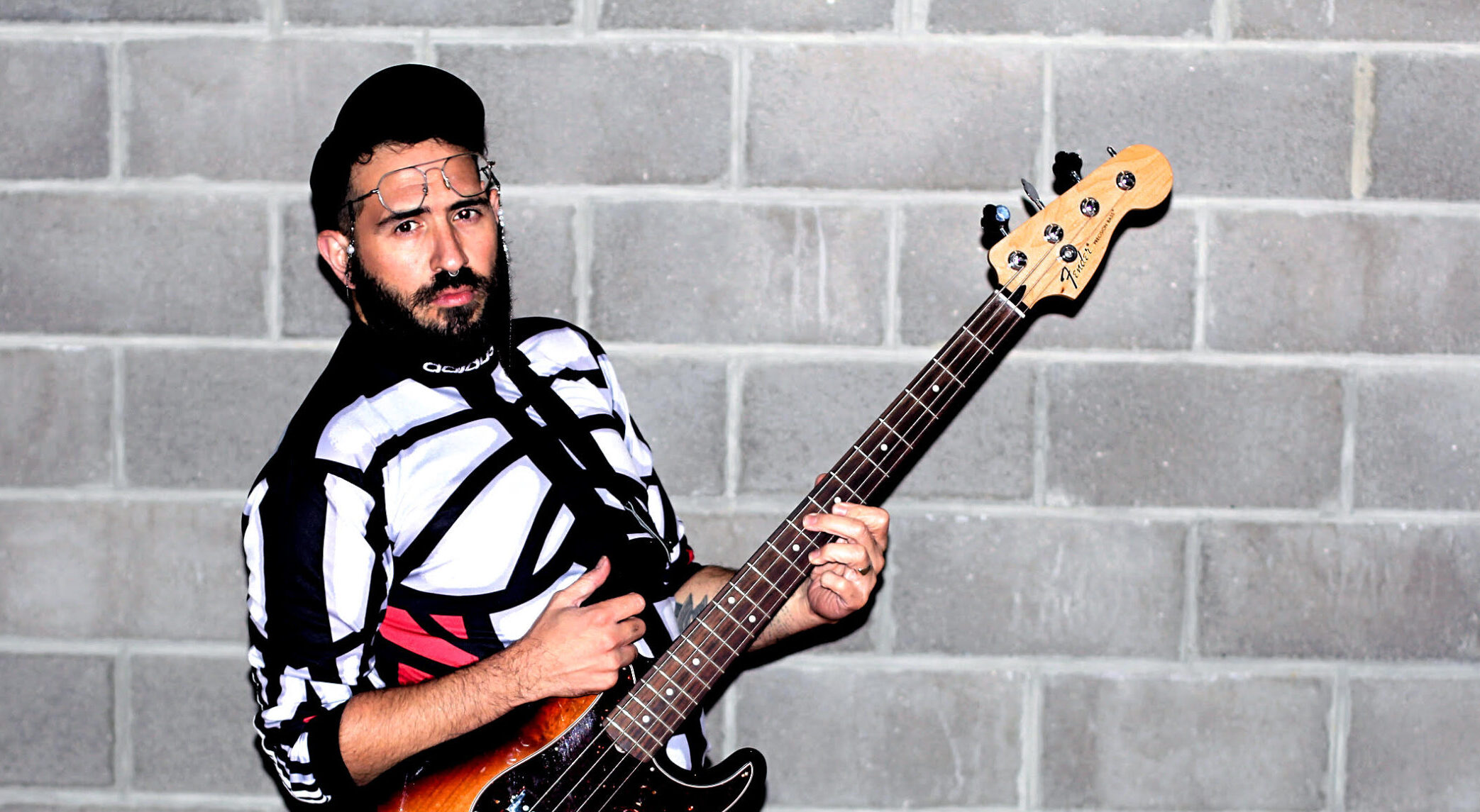 A white person with a dark beard wears glasses, a black baseball cap, a patterned skirt and a black and white sweater. They hold an electric guitar. 