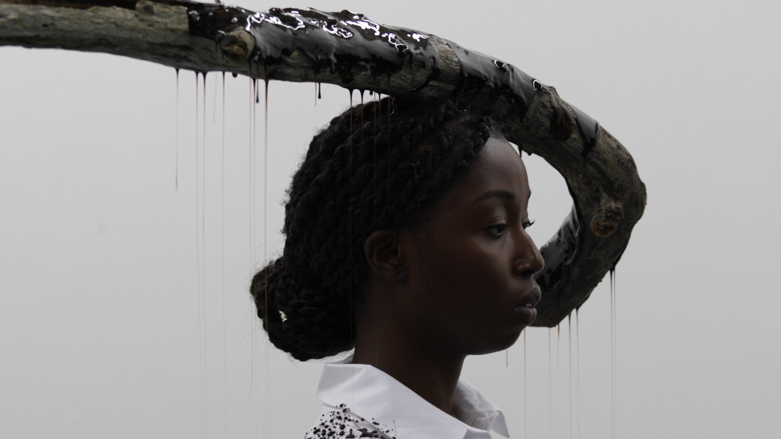 A black woman with her hair in a bun stands infront of a tree branch, which is covered in sticky black fluid. 