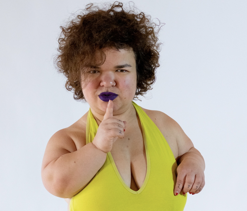 Artist Tammy Reynolds wears a yellow swimsuit and black lipstick. They are holding their finger to their mouth in a shhh gesture.