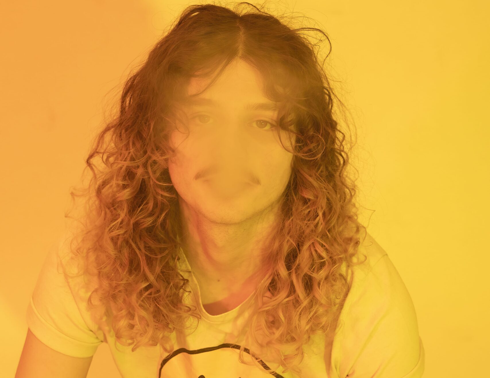 A white man with a moustache and curly blonde-brown hair sits on the floor, wearing a white tshirt with a smiley face. The image is tinted yellow. 