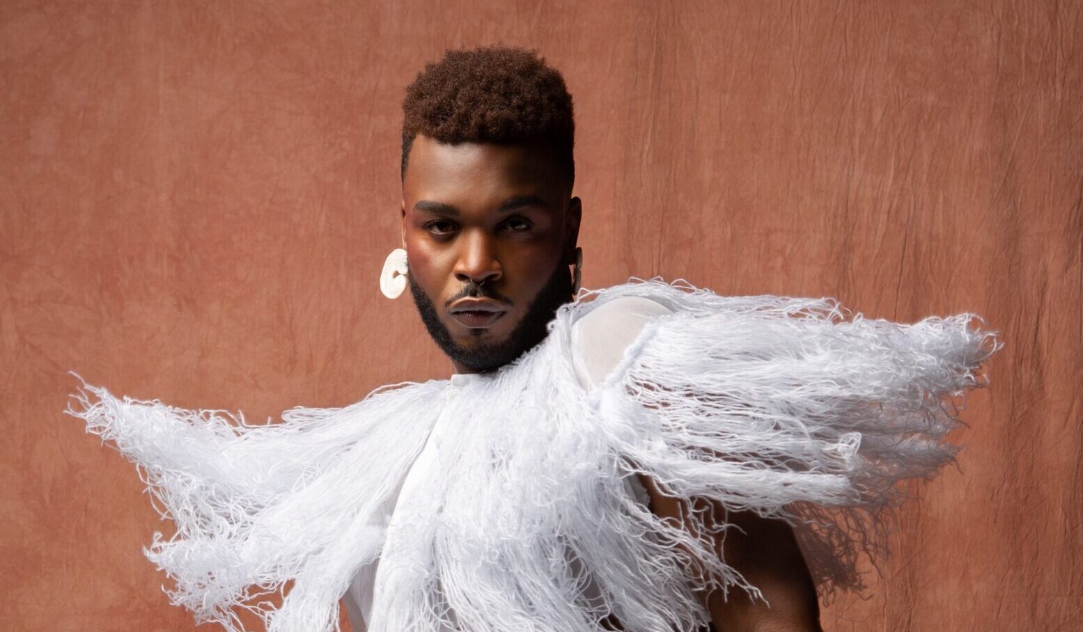 Cakes Da Killer wears a white fringed cape and skirt, and a white hoop earring.