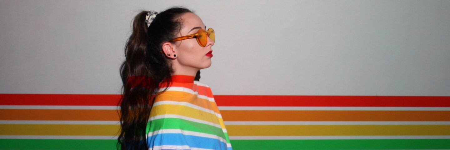 A white woman with long brown hair and sunglasses stands in a projection of rainbow lines. 