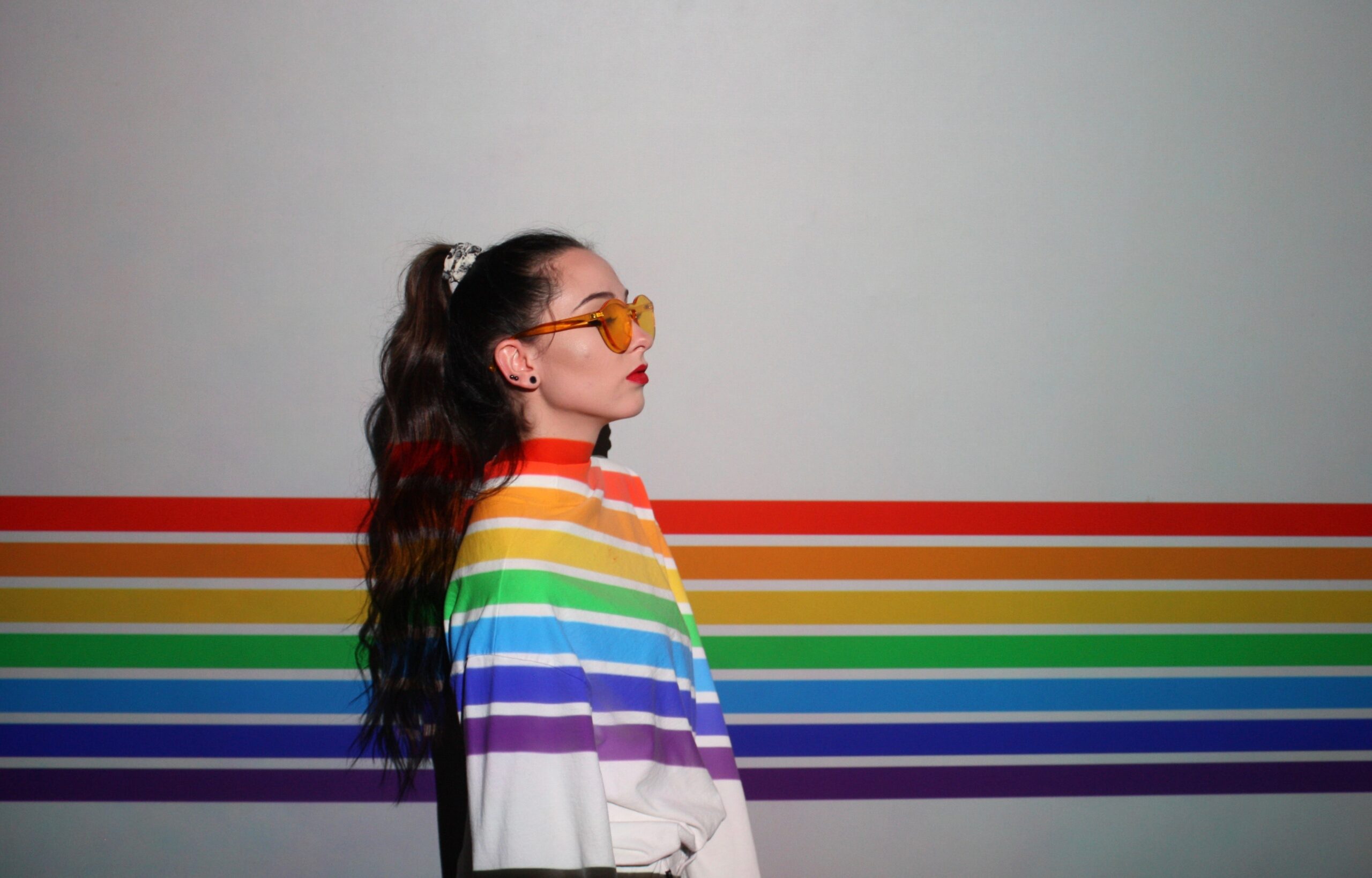 A white woman with long brown hair and sunglasses stands in a projection of rainbow lines. 
