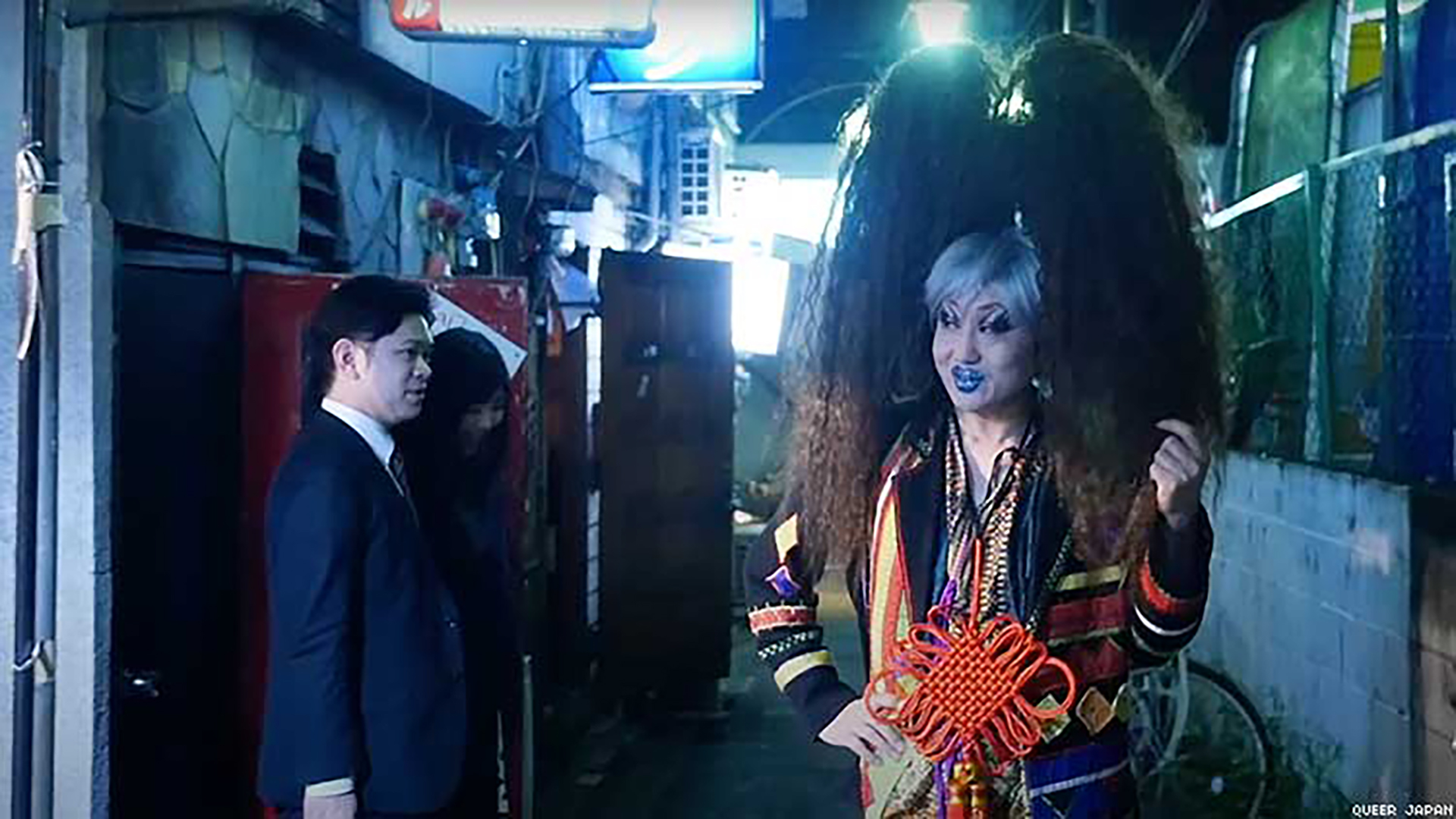A Japanese drag artist in a huge wig smiles at a businessman in a back alley.