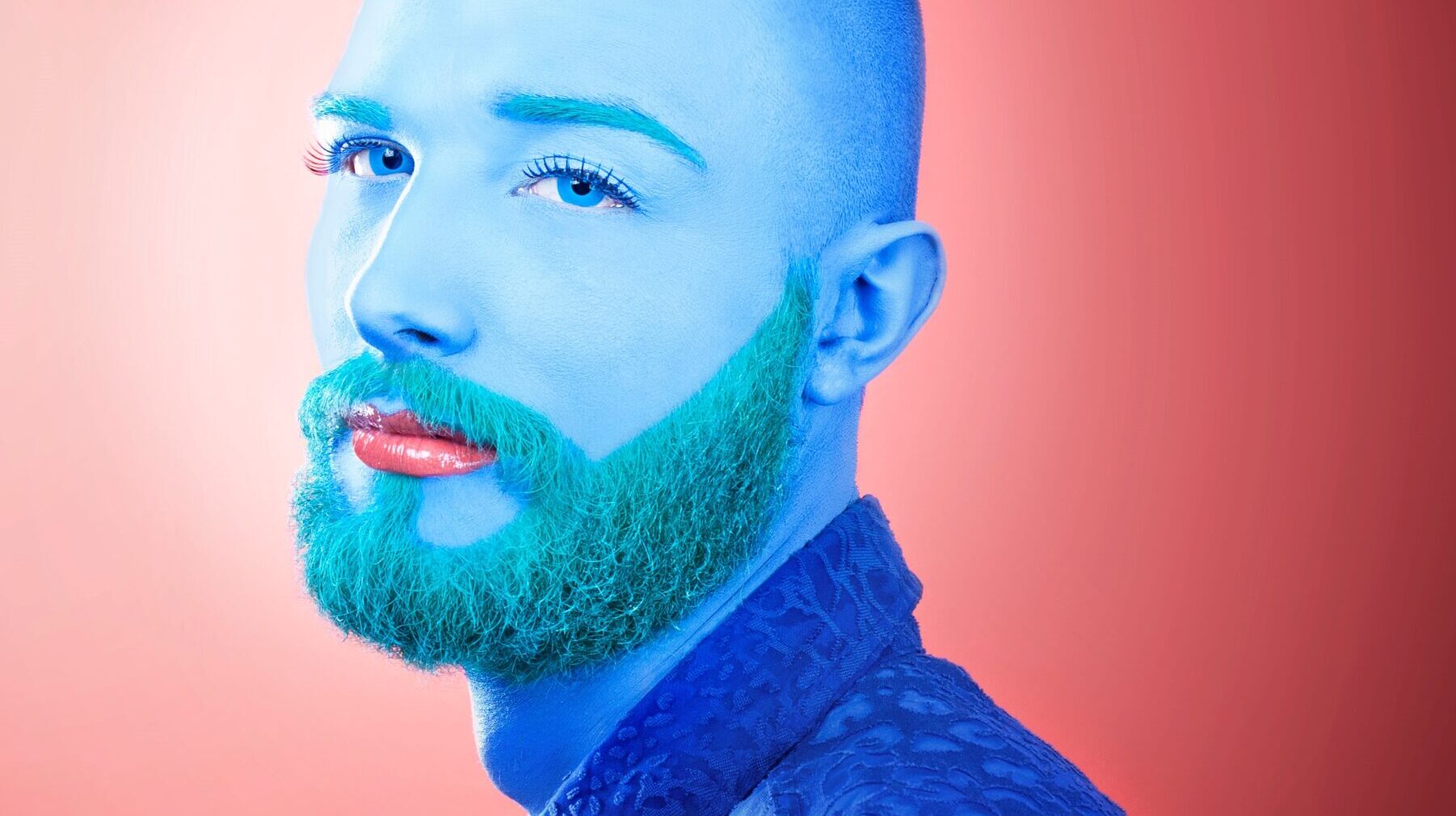 A white man in a blue shirt sits infront of a red background. He face and beard are painted blue, and he has pink lipstick on.