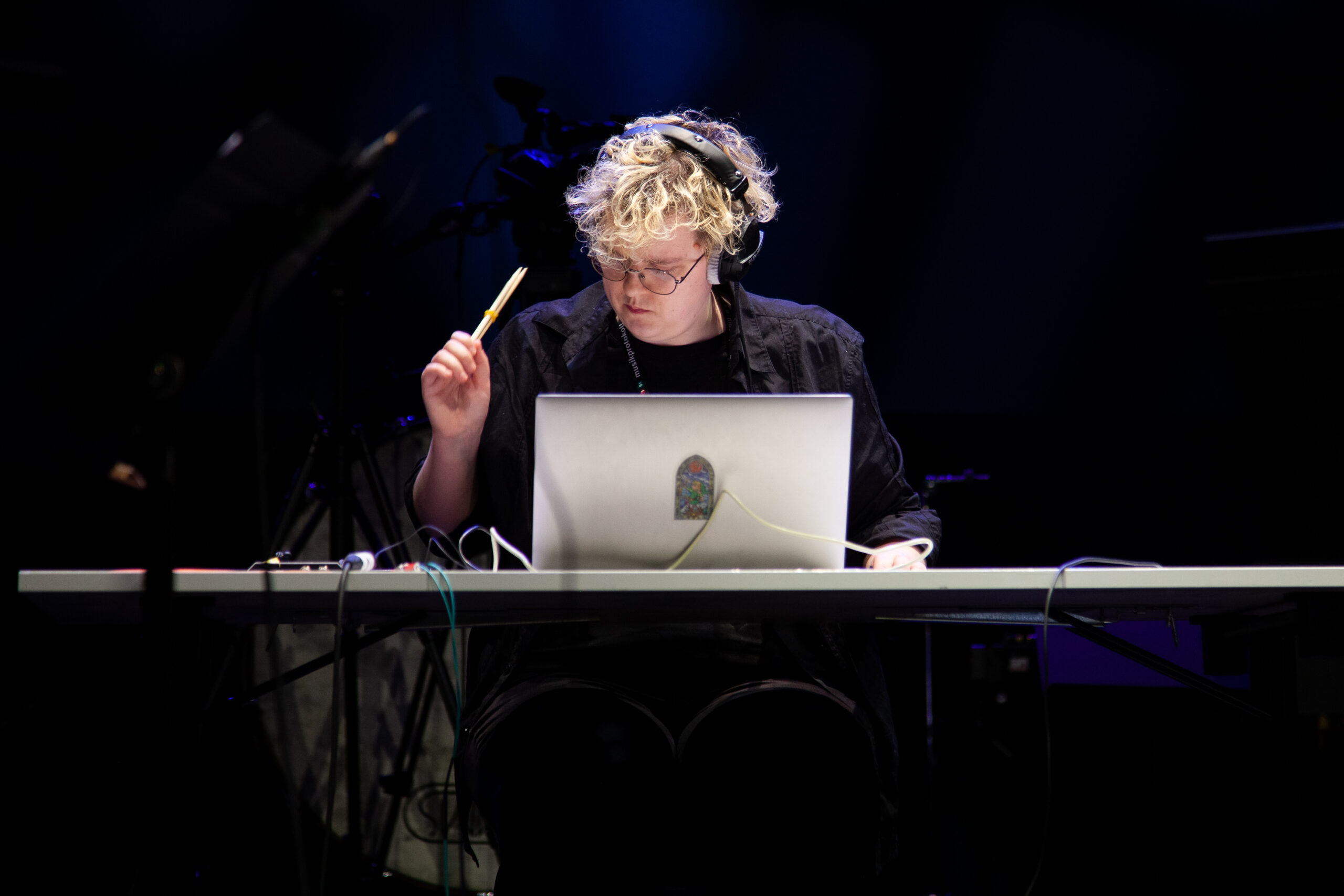 A person sits at a desk with a silver laptop. They have blond wavy short hair and black headphones on. There are wires lying on 