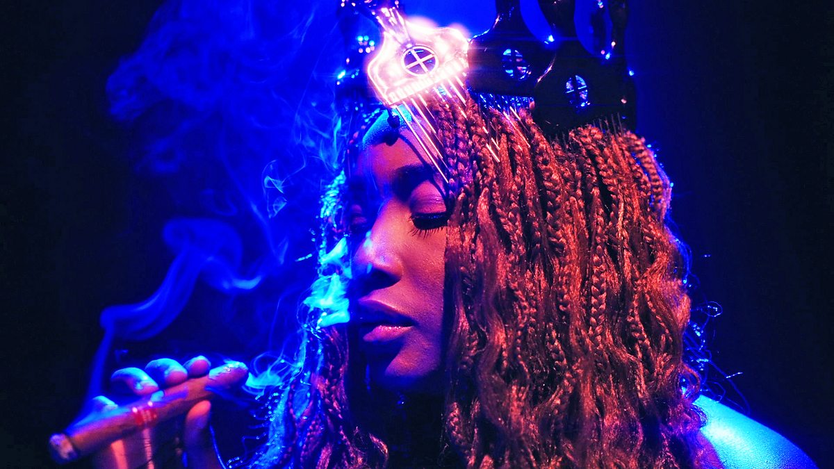 A Black woman with braided hair smokes a cigar in blue light.