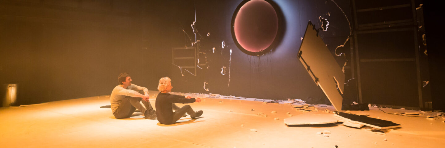 Two white people sit looking at a destroyed black plasterboard wall as part of a performance. 