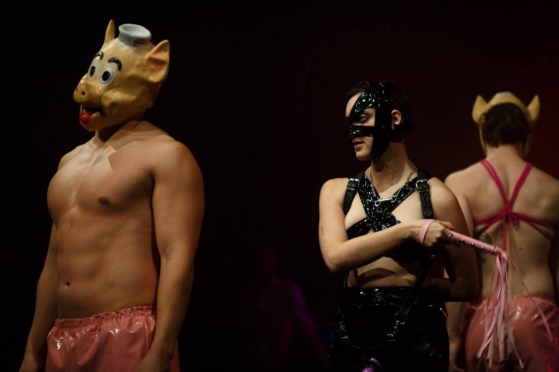A white person in black latex holds a whip. There are also two white people in pig masks and pink latex. 